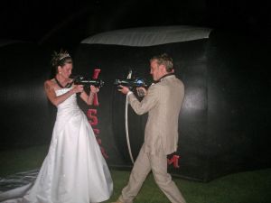 A Bride and Groom playing laser tag on their wedding day. Both are standing outside the laser tag dome and shooting each others laser guns.