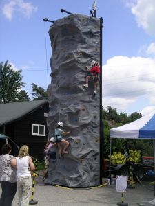 Mobile rock climbing wall being climbed by children while two adults watch them climb the wall.