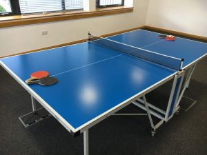 Ping pong table set up in a conference room at a office complex. The blue table tennis game has four table tennis bats and balls on the playing surface.