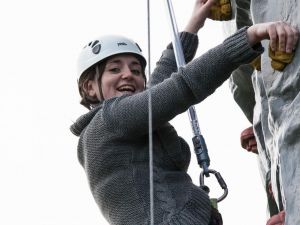 A happy female climbing a mobile rock climbing tower while wearing a rock climbing harness and helmet.