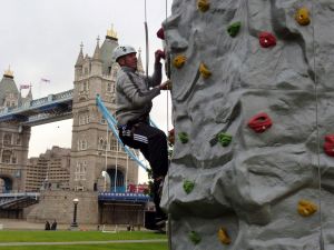 Rock climber ascending the mobile rock climbing wall at an event next to Tower Bridge in Central London. The mobile climbing wall is on the banks of the River Thames with a male climber wearing a harness and rock climbing helmet.